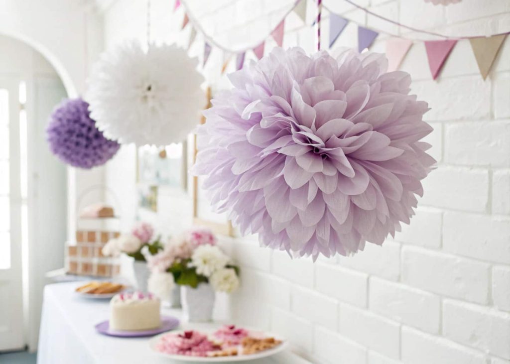 A large fluffy tissue paper pom-pom flower in soft lavender and white tones, hanging against a white wall, festive party decor style, bright and airy, shallow depth of field.