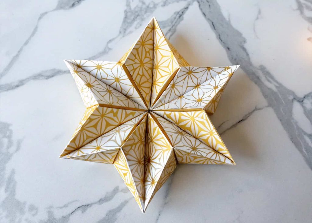 A geometric star-folded origami flower in gold and white patterned paper, placed on a marble surface, flat lay photography, crisp and modern aesthetic, bright overhead lighting.