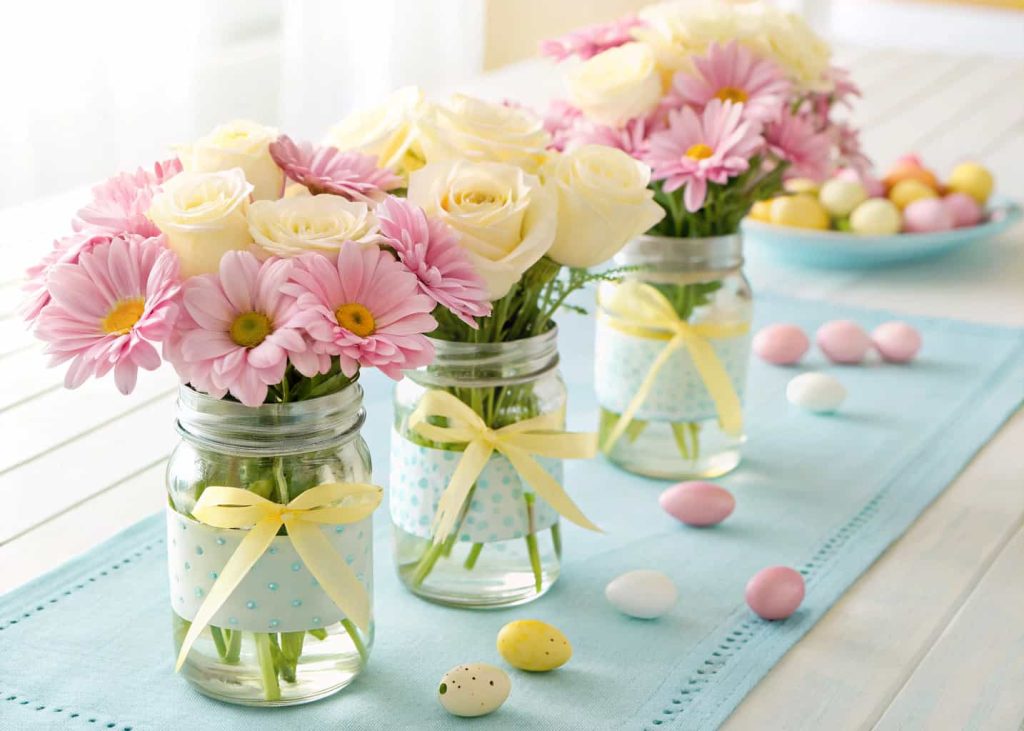 Three mason jars filled with pink daisies and white baby roses, tied with pastel yellow ribbon, arranged in a row on a light blue table runner, small colored Easter eggs nearby, bright morning light