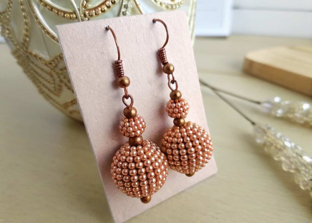 A pair of handmade beaded earrings crafted from acrylic beads painted with rose gold metallic nail polish hanging on a small display card