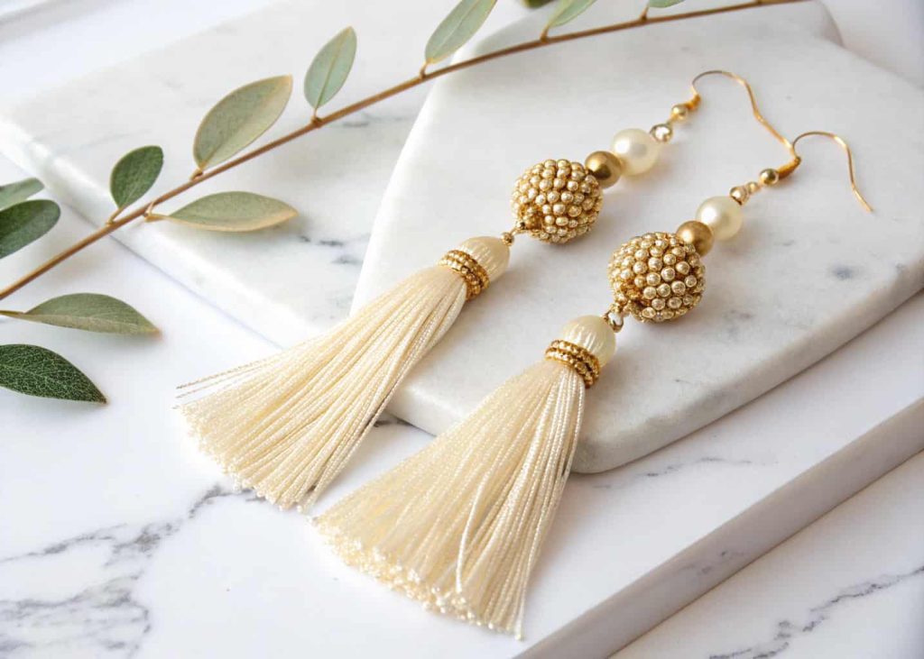 Handmade gold bead and cream tassel earrings displayed on a marble surface with soft natural light showing elegant movement and texture