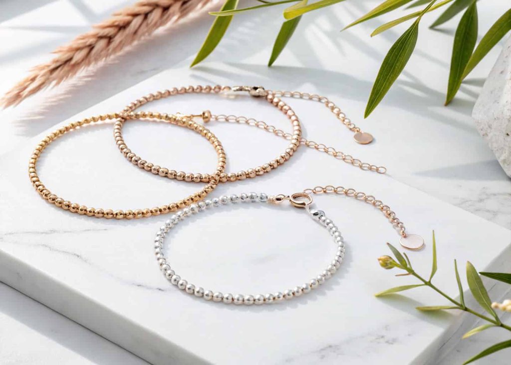 A minimalist jewelry flat lay showing three thin bracelets with tiny beads in gold, silver, and rose gold tones, arranged parallel on a clean white marble surface, overhead shot, bright natural lighting.