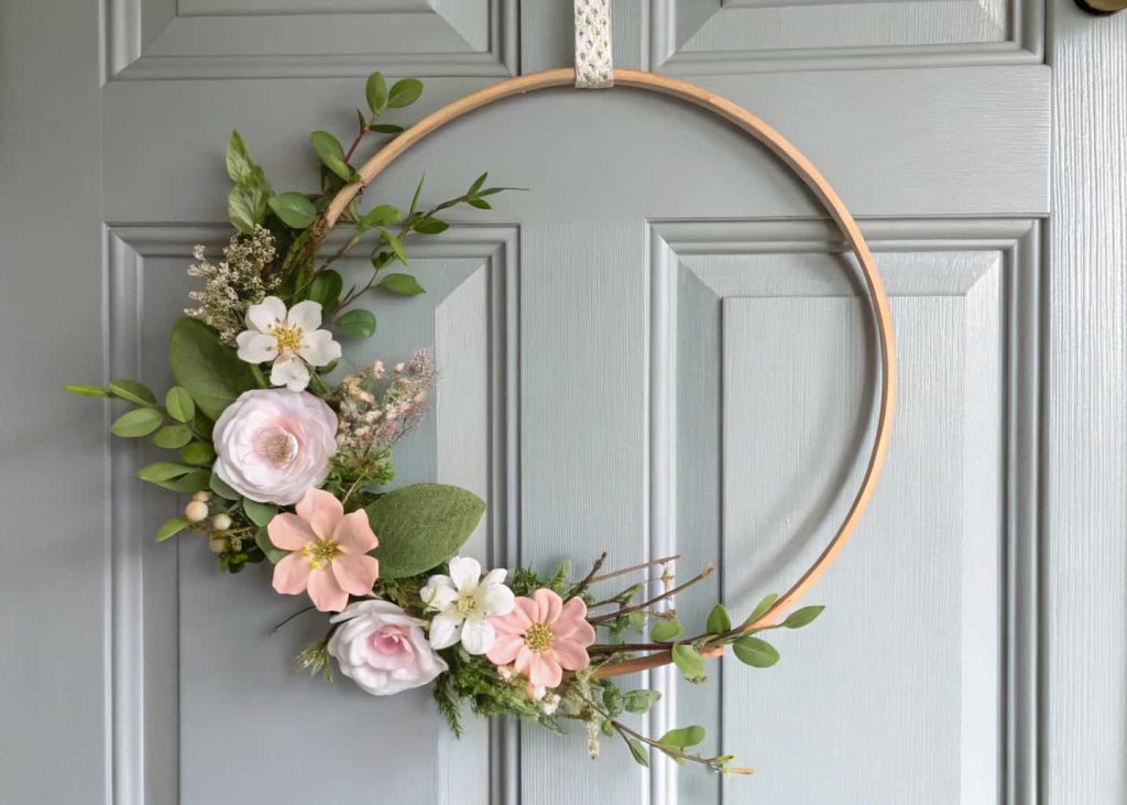 A minimalist spring wreath with a thin wooden hoop, a few pastel flowers, and green leaves on a modern gray door.