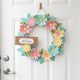 A bright, floral spring door hanger on a white front door with pastel-colored flowers and a small welcome sign.