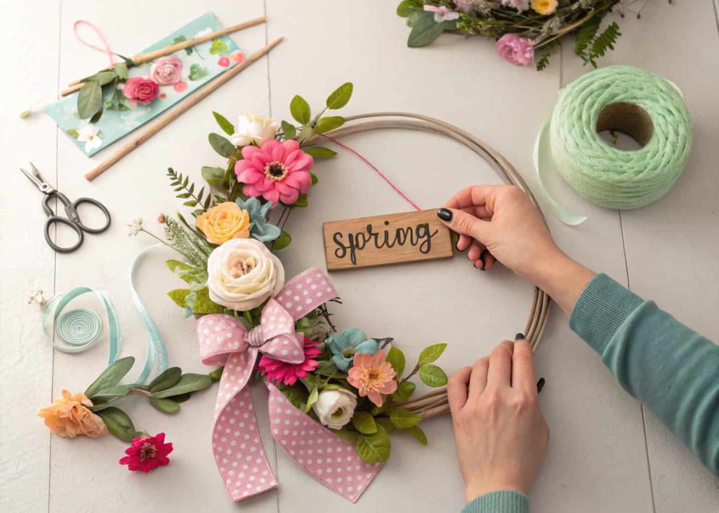 Hands crafting a spring door hanger with faux flowers, ribbons, and a small wooden sign on a table.