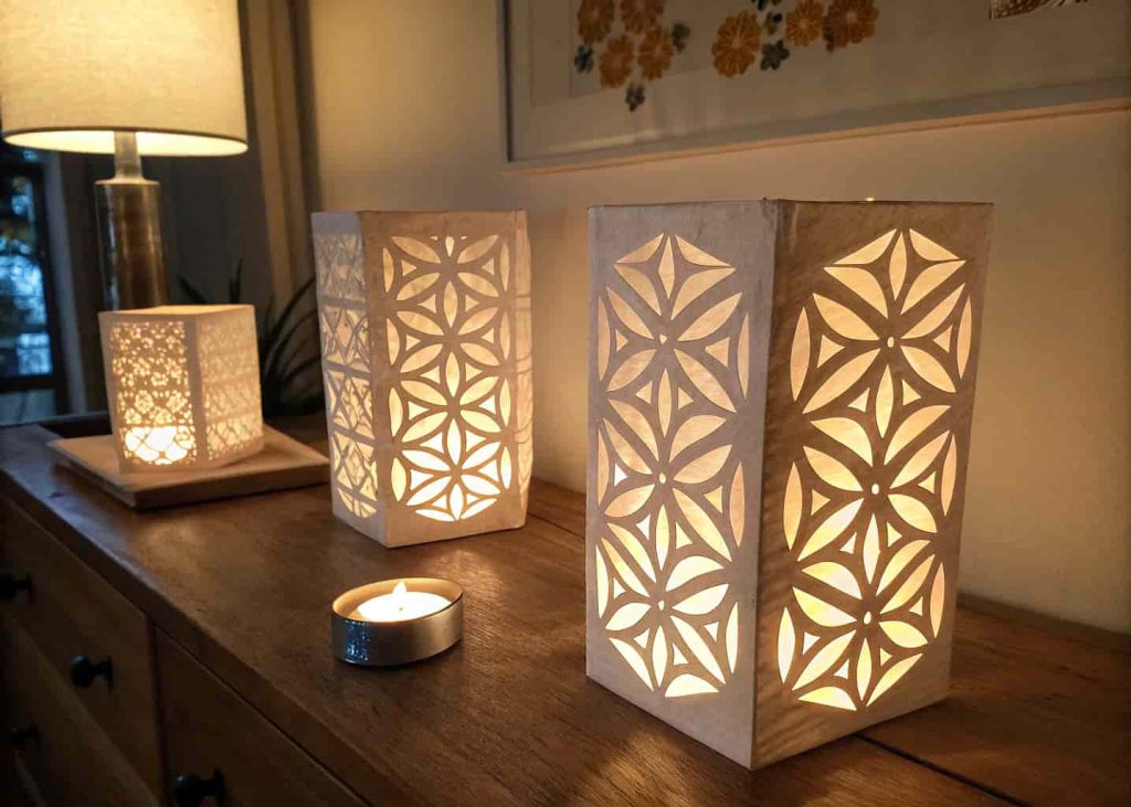 Handmade paper lanterns with geometric cut-out patterns glowing softly on a wooden surface in dim warm lighting.