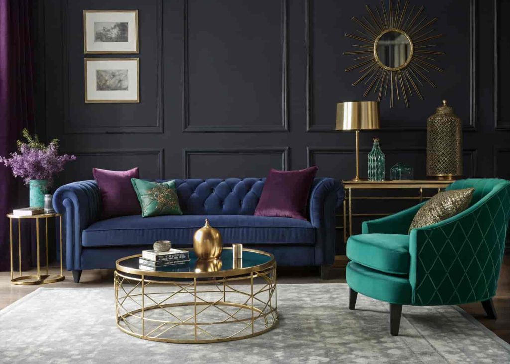 Art Deco living room with sapphire blue sofa, amethyst pillows, emerald green chair, and gold coffee table in a moody setting.