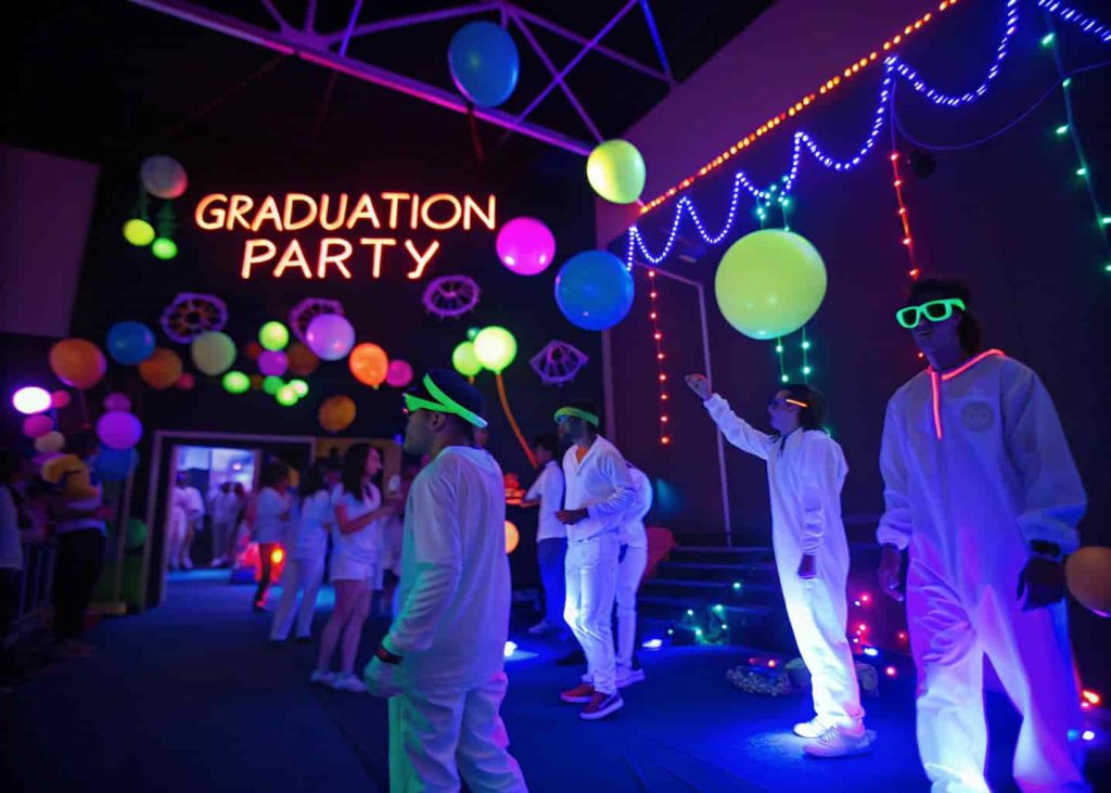 Indoor graduation neon glow party with UV black lights, guests in glowing white outfits, neon balloons, and colorful glowing decorations.
