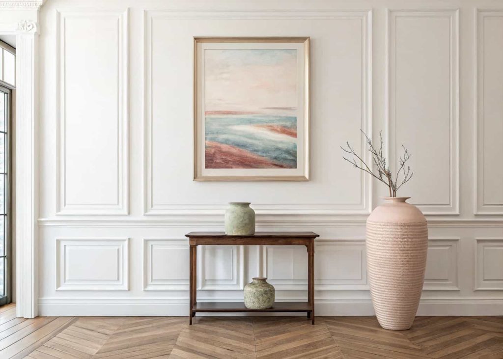 A minimalist wall with one large framed abstract art piece in soft tones, a single tall ceramic vase below it on a console table, natural wood floor, and clean white walls — intentional, gallery-like, and quietly elegant.