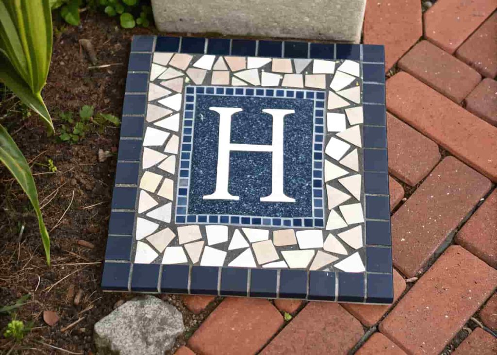 Personalized mosaic stepping stone with the letter H in white tiles on a dark blue background in a garden.