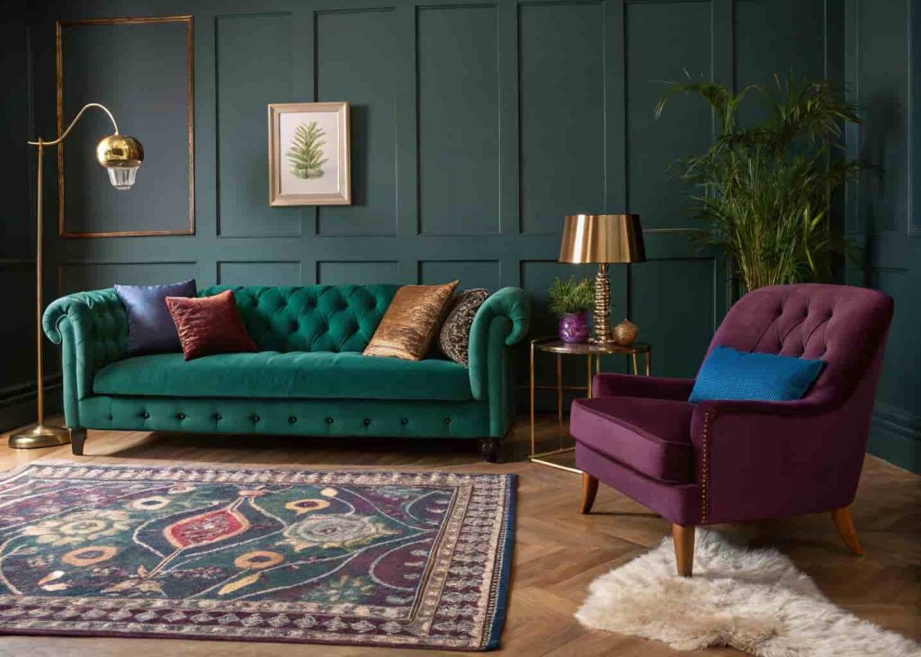 A jewel-toned maximalist living room with emerald green sofa, sapphire armchair, amber rug, and brass accessories glowing under warm ambient lighting.