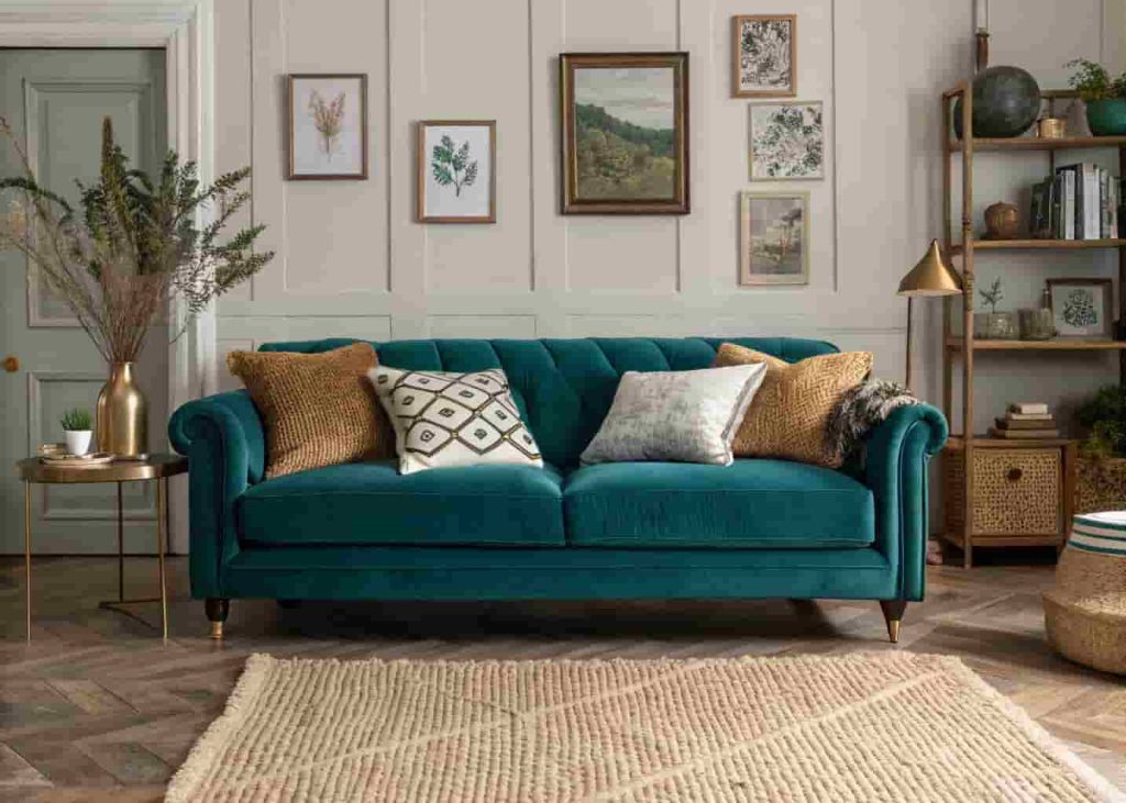 A deep teal velvet sofa in a maximalist living room styled with linen and chunky knit cushions, a jute rug, and warm brass accessories.