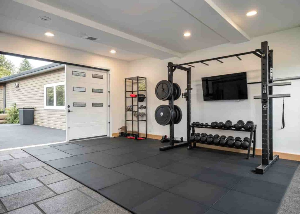 Finished garage home gym with rubber floor tiles, squat rack, dumbbells, and bright lighting