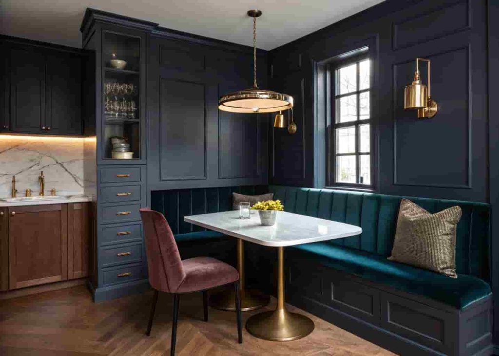 Dark moody breakfast nook with velvet bench, navy walls, marble table, brass pendant light, and hidden storage in a dramatic kitchen corner