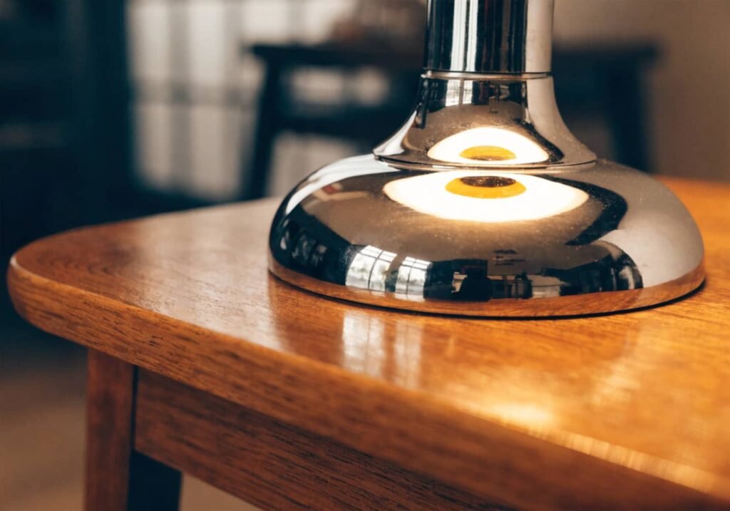 A real photo close-up of a chrome table lamp base reflecting warm indoor lighting, set on a wooden side table next to a small potted plant
