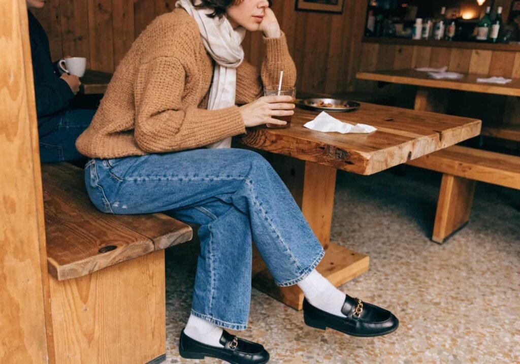 Person in a cozy caramel knit sweater and scarf sitting at a rustic cafe table.