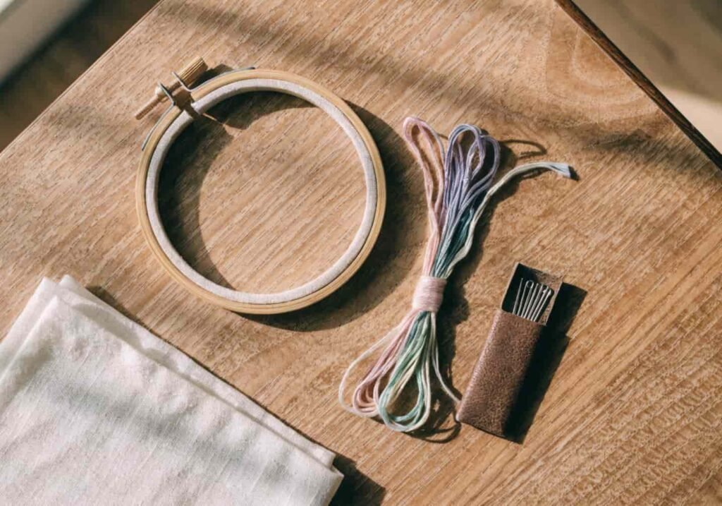 Flat lay of embroidery supplies including hoop, pastel floss, needle, and cotton fabric
