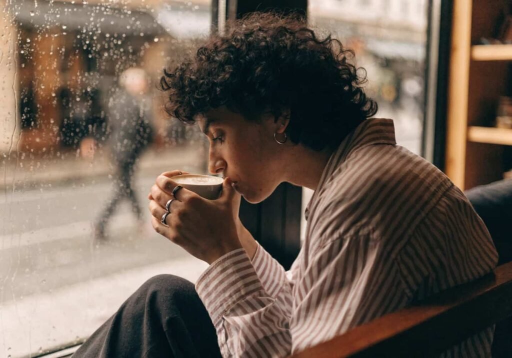 Person sitting by a rainy cafe window holding a coffee cup with soft natural light