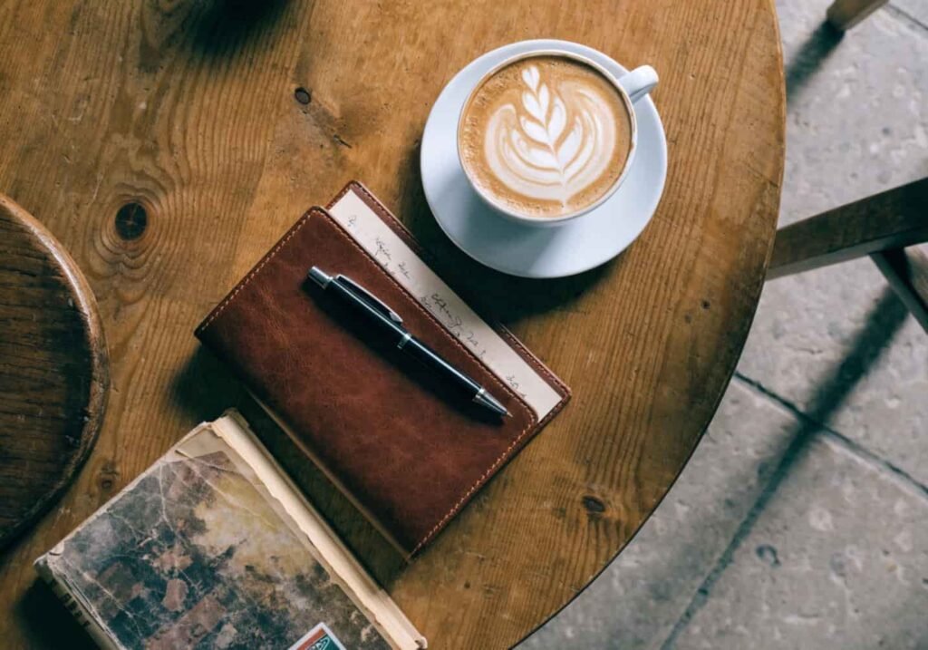 Flat lay of latte art coffee, journal, and book styled on a wooden cafe table.