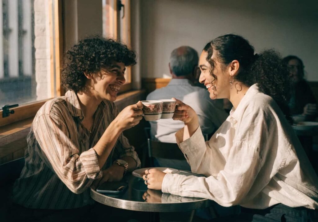 Two friends clinking coffee cups together and smiling at a cafe table