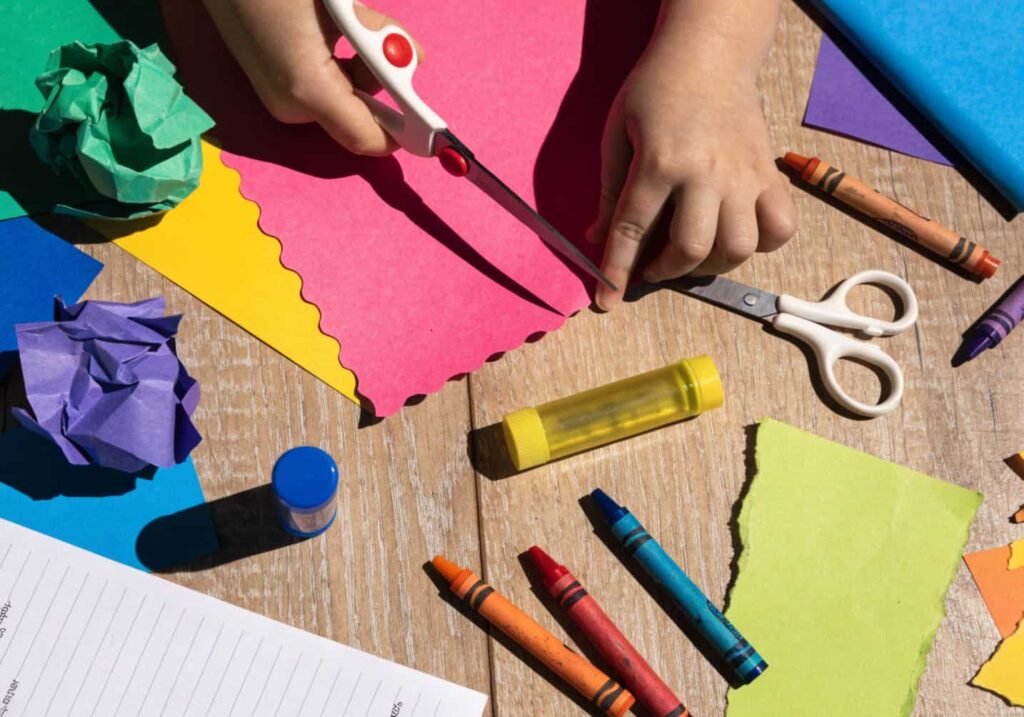 Kindergarten child practicing scissor skills with construction paper and craft supplies on a table.