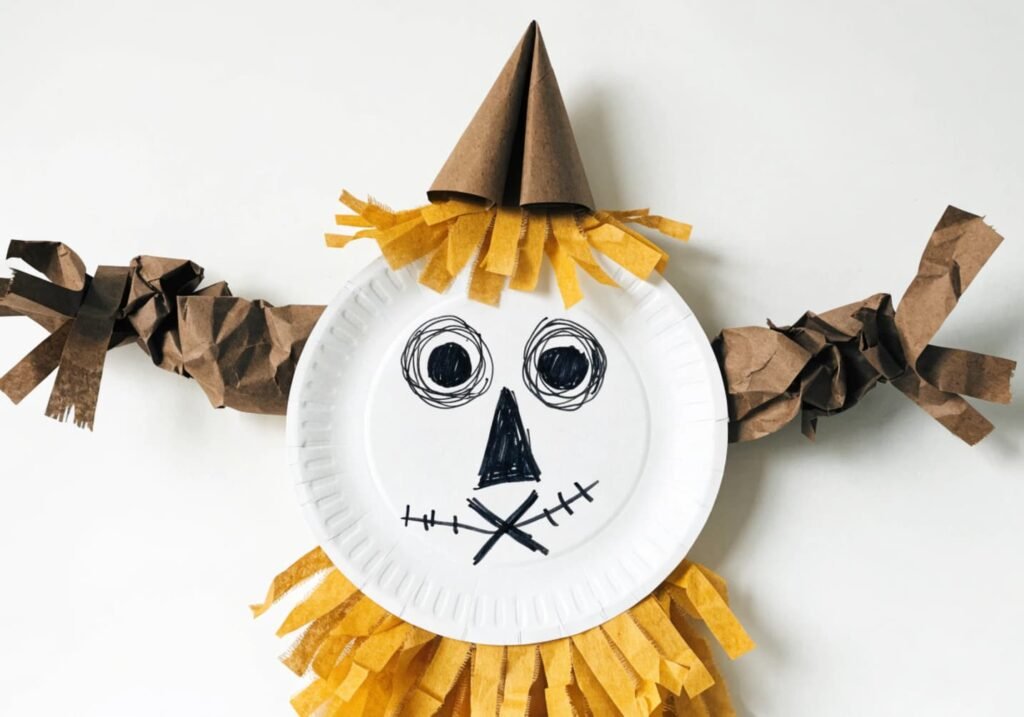 Paper plate scarecrow craft with a stitched smile and crepe paper straw trim.