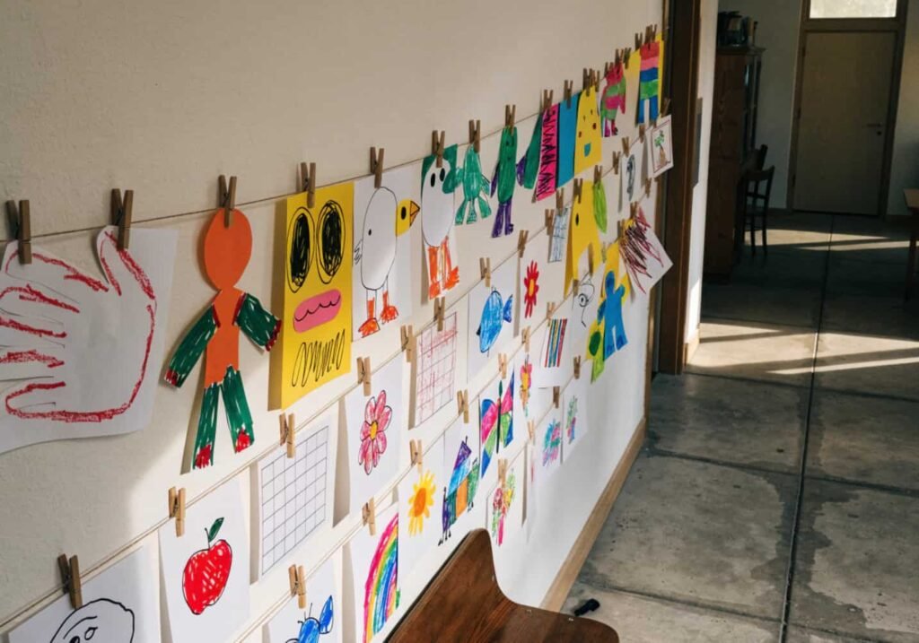 Kindergarten artwork displayed on a clothesline with wooden clips along a classroom wall.