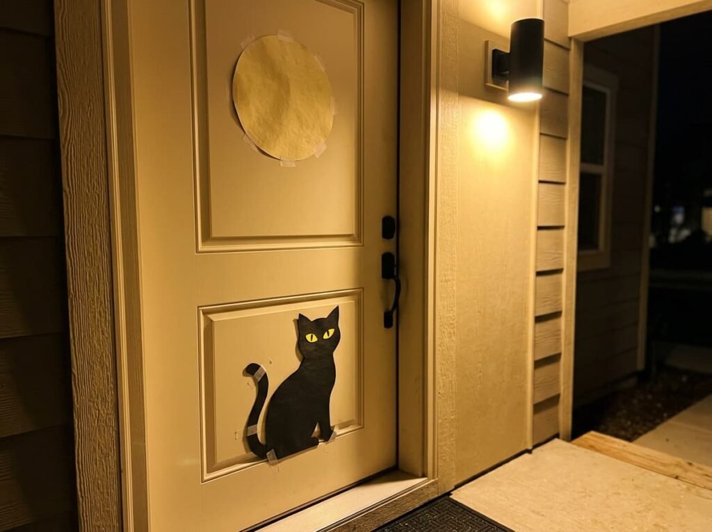 Black cat silhouette with yellow eyes on a cream front door under a paper moon